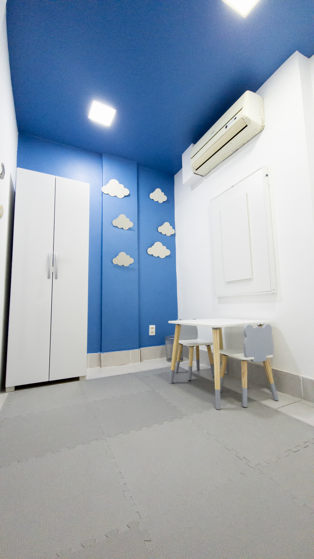 Sala infantil com parede azul, nuvens decorativas e mesinha com cadeiras para crianças.