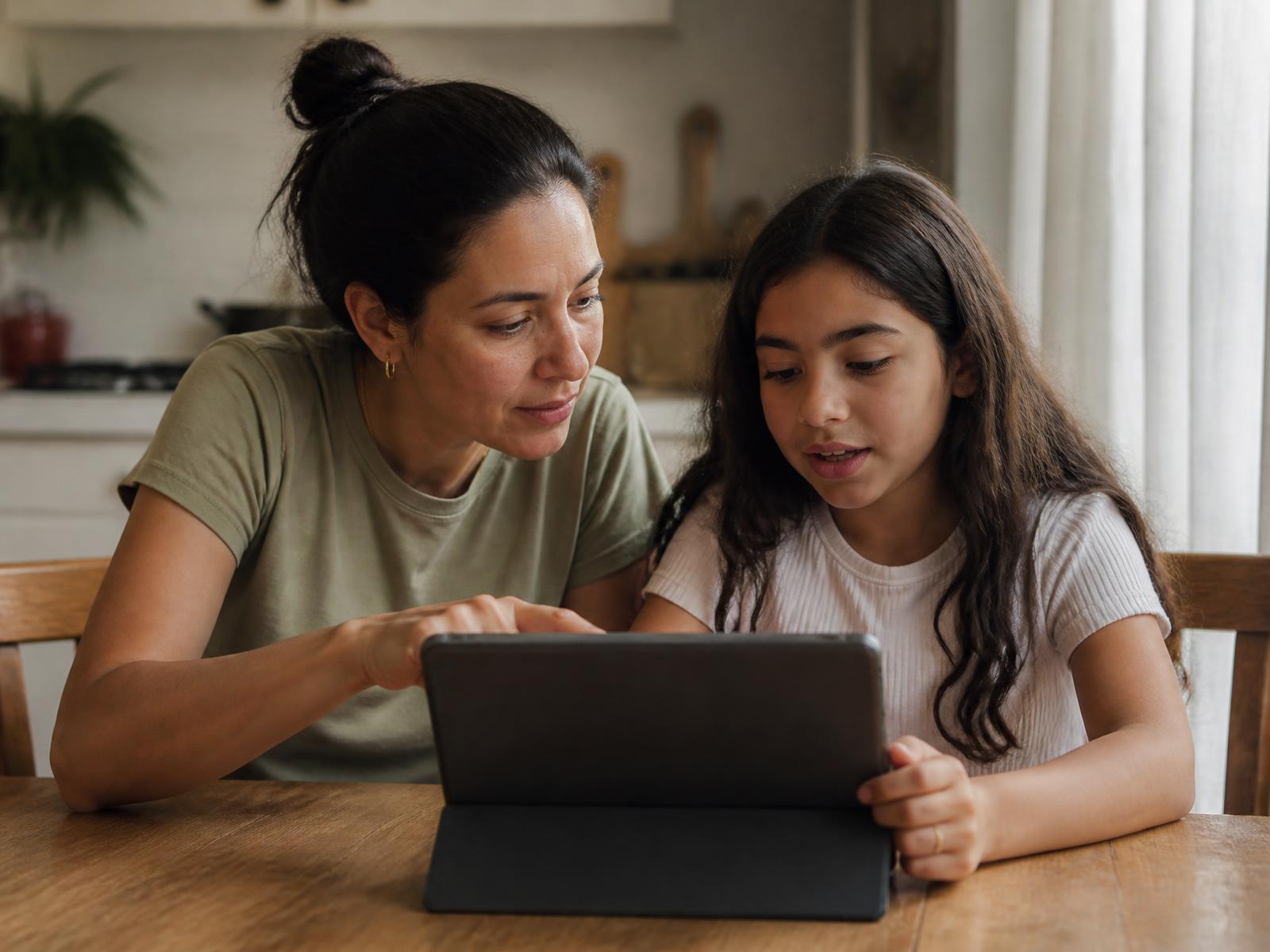 Mãe e filha sentadas à mesa da cozinha olhando juntas a tela de um tablet