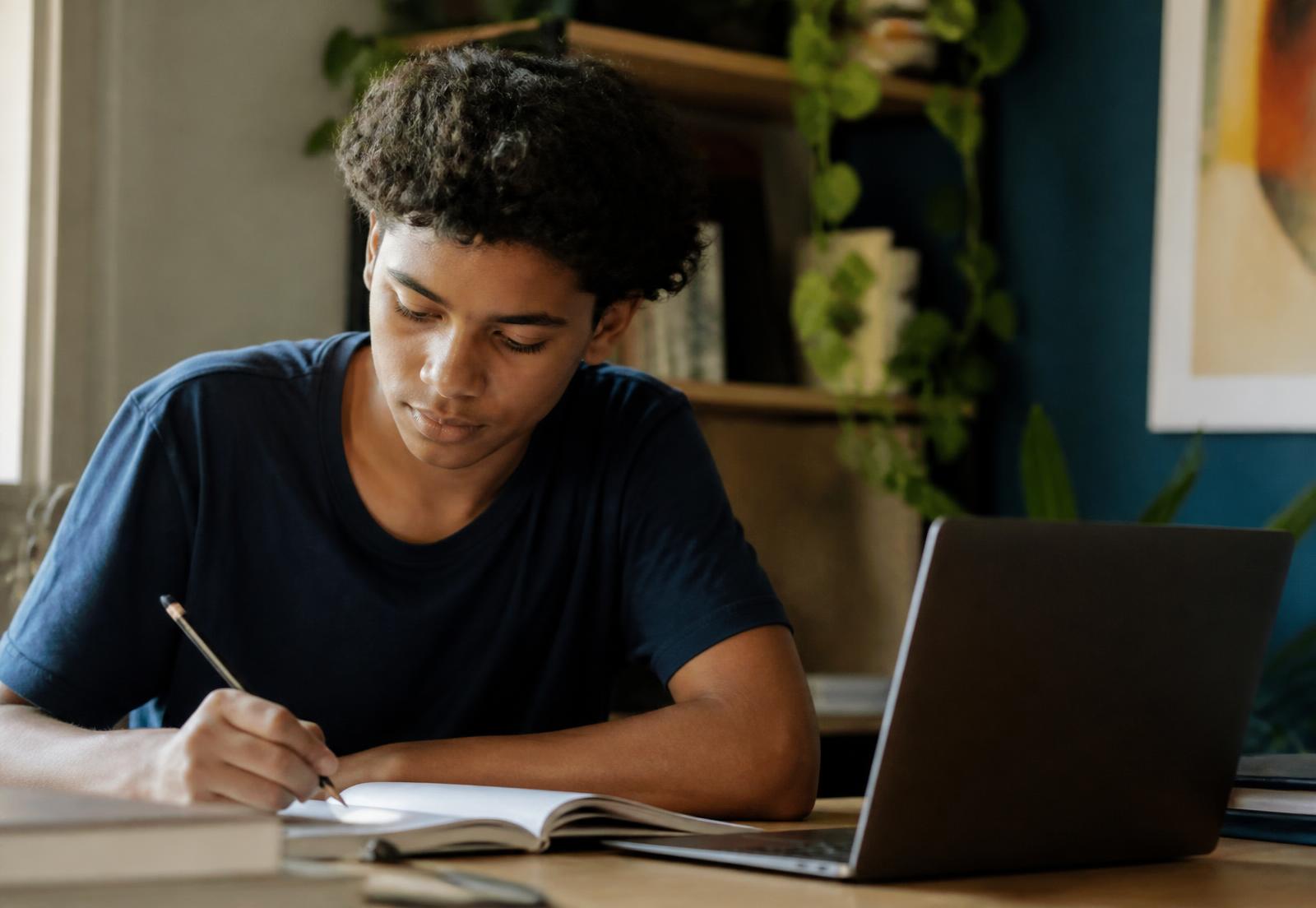 Adolescente estudando com caderno aberto e um notebook parcialmente fechado ao lado
